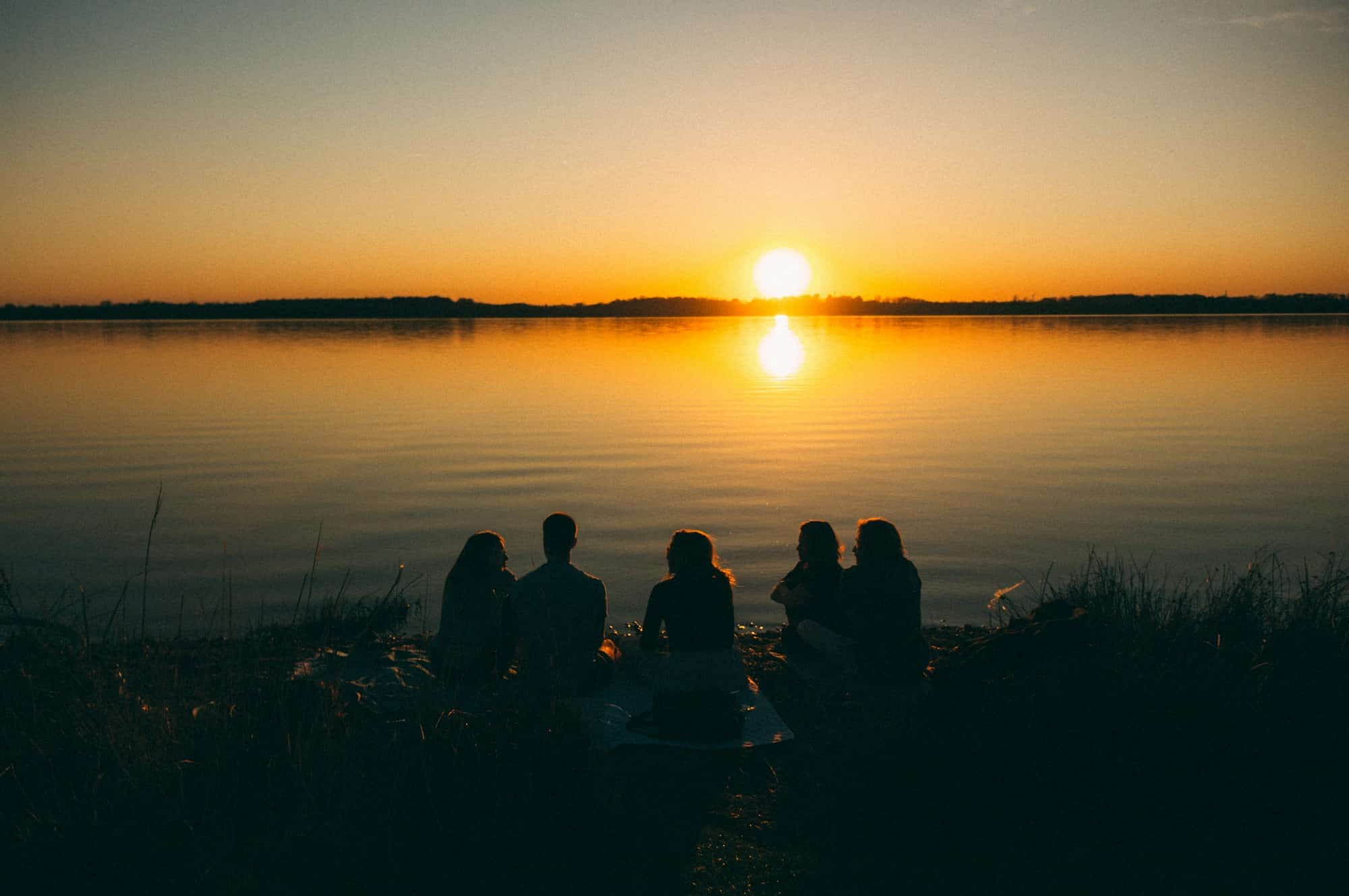 Fünf junge Leute sitzen als Silhouetten am Ufer des Cospudener Sees vor dem Sonnenuntergang