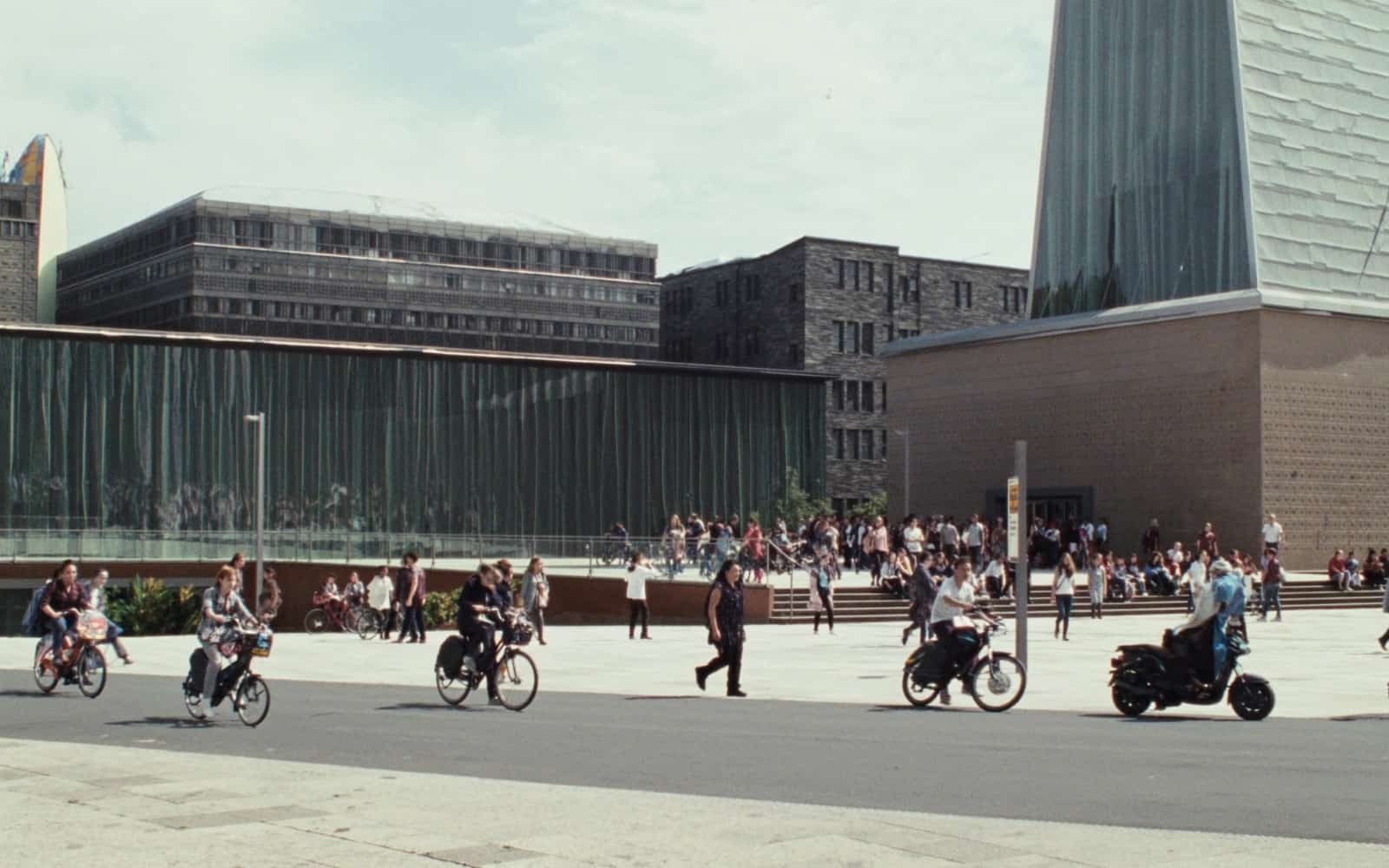 Belebter Platz in Leipzig mit jungen Menschen auf Fahrrädern und Gruppen auf Sitzstufen vor moderner Architektur